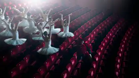 En grupp ballerinor springer mellan rader av röda stolar.
