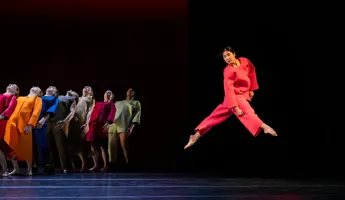 A group of dancers in colorful clothes, in the foreground a dancer in red jumps.