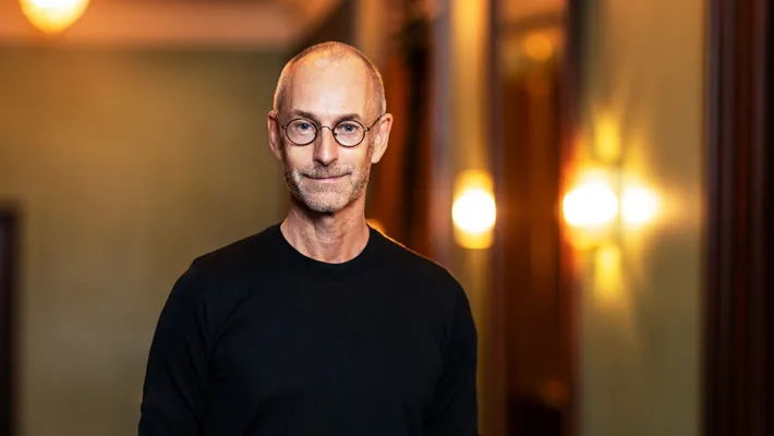 A smiling man in a black shirt and glasses against a blurred background with multiple light sources.