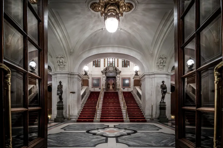 Utsikt över en stor entréhall, en trappa med röd matta leder uppåt mot en dörr.