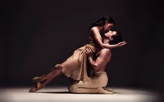 Two ballet dancers in a dynamic pose on a stage with a dark background, emphasizing their movement and expression.