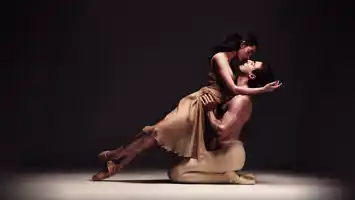 Two ballet dancers in a dynamic pose on a stage with a dark background, emphasizing their movement and expression.