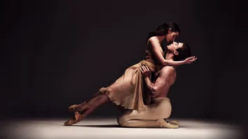 Two ballet dancers in a dynamic pose on a stage with a dark background, emphasizing their movement and expression.