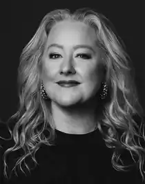 A close-up of a blonde woman wearing a dark sweater and large earrings.