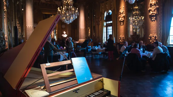 In the foreground a harpsichord, in the background several set tables with guests.