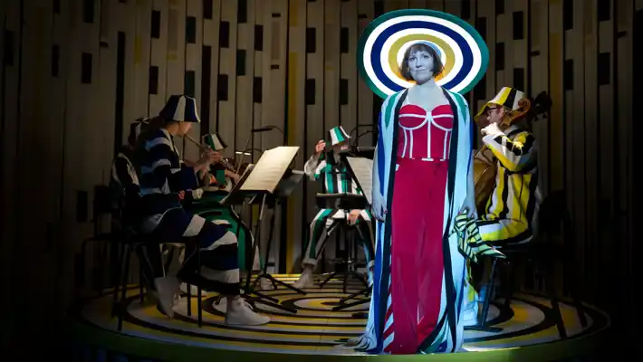 A woman with a large hat stands in front of an orchestra and looks into the distance.