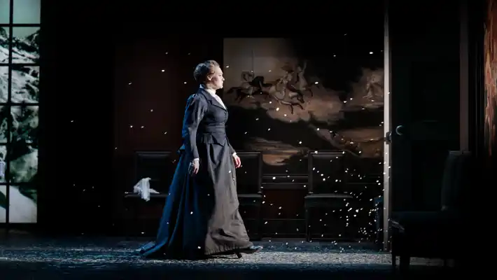 A woman in a dark, long dress stands in the light of a doorway, snow swirling around her.