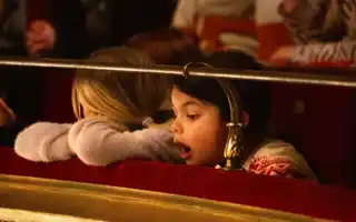 Person with blond hair rests his head on the ledge in a theater - a second person looks open-mouthed towards the stage.
