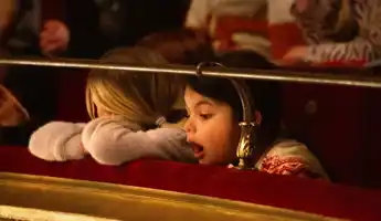 Person with blond hair rests his head on the ledge in a theater - a second person looks open-mouthed towards the stage.