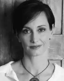 A close-up of a woman with dark short hair, she is wearing a white shirt and has a large necklace.