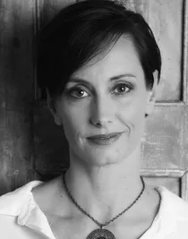 A close-up of a woman with dark short hair, she is wearing a white shirt and has a large necklace.