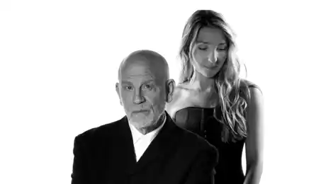 A man in a suit, behind him stands a woman with long hair.