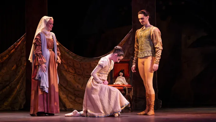 A woman kneels before a man, behind her stands another woman who appears older.