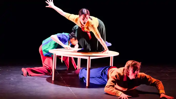 Three dancers move over, under and behind a table in colorful costumes.