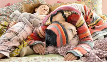 Two people lie down and sleep, blankets, pillows and stuffed animals are with them in bed.