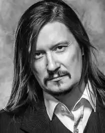 A close-up of a long-haired man with a mustache, wearing a light shirt and suit jacket.