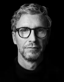 A person wearing a dark sweater stands against a black background. The face is partially in the dark, making it difficult to distinguish details.