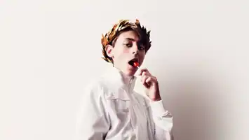 Young person in a white shirt with a laurel wreath on his head and a lollipop in his mouth against a light background.