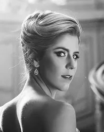 A close-up of a blonde woman, her back turned to the camera, she looks over her shoulder at us.