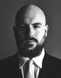 A close-up of a man, he has a beard and mustache, is dressed in a suit and wears a tie.