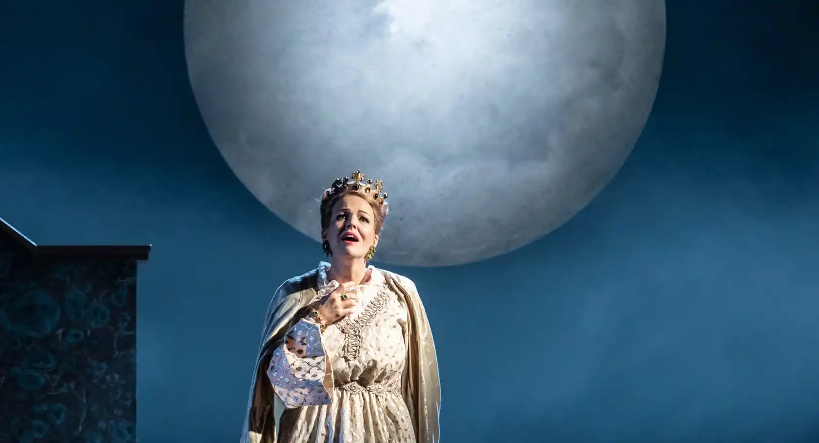 A woman in a cloak and with a crown on her head, in the background a large moon.
