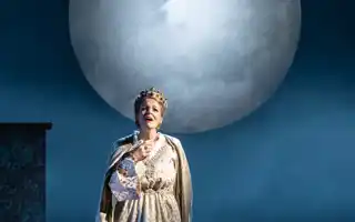 A woman in a cloak and with a crown on her head, in the background a large moon.