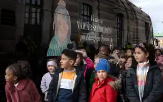 A group of children of various ages wait in front of a bus on the sidewalk.