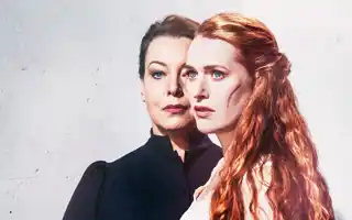 Two women against a light background, in the foreground a red-haired woman with a scar on her cheek.