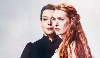 Two women against a light background, in the foreground a red-haired woman with a scar on her cheek.