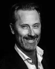 A close-up of a smiling man, he is wearing a dark suit and has a light unbuttoned shirt.