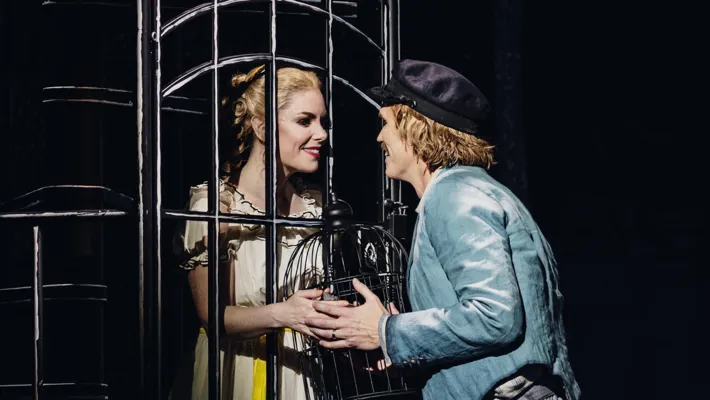 A woman stands behind a grate, a man on the other side, they hold a cage.