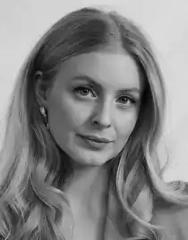 A close-up of a woman with long blonde hair: she is wearing a light jacket.