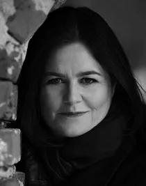 A close-up of a woman in dark clothes, standing partly behind a wall on the left side.