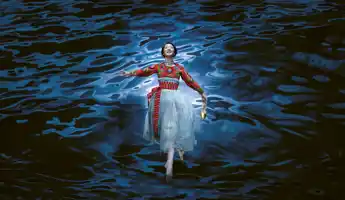 A woman, wearing a tulle skirt and folkloric jacket, floats over blue water: in her hand she holds a golden shoe.