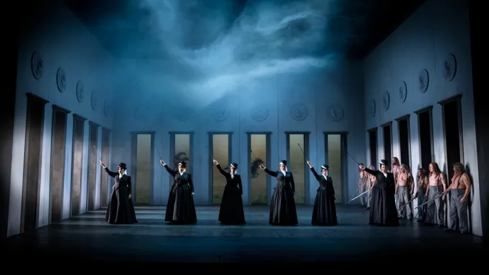 Six women in black clothes stand in a room with many doors, with riding sticks in their hands, stretched upwards.
