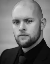 Close-up of a young bearded man wearing a dark suit and dark tie.