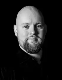 Close-up of a young bearded man wearing a dark suit and dark tie.