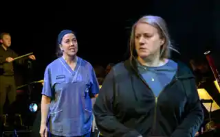 A woman in hospital clothes stands diagonally behind another woman, in regular clothes.