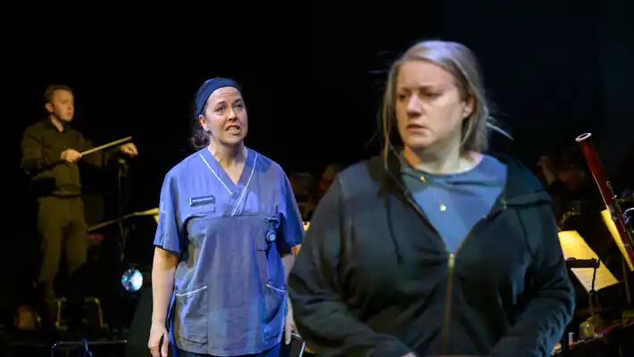 A woman in hospital clothes stands diagonally behind another woman, in regular clothes.