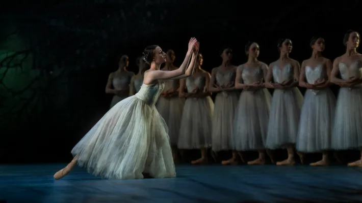 A ballerina in a long white tulle skirt stands with her leg outstretched, behind her are several female dancers in white.