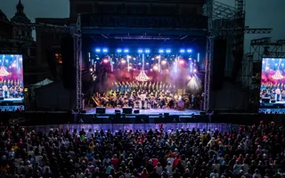 A large crowd of people in front of a lit stage, with two large video screens on the sides.