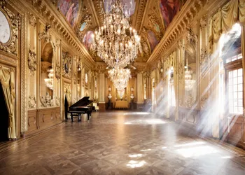 A gold-decorated room with a grand piano on the left, the sun shining through windows on the right.