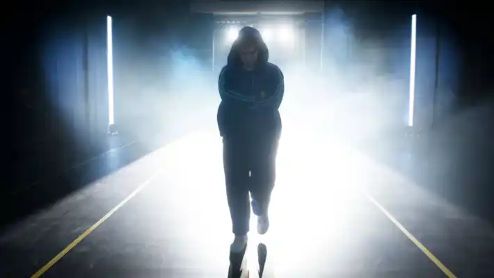 A boy with his arms crossed and a drooping gaze stands facing the camera with bright backlight in the background.