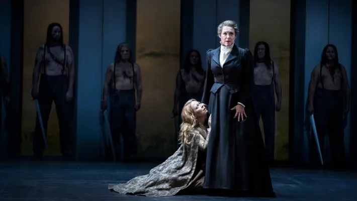 A woman is half-lying on the floor, reaching up towards another woman, pleading.