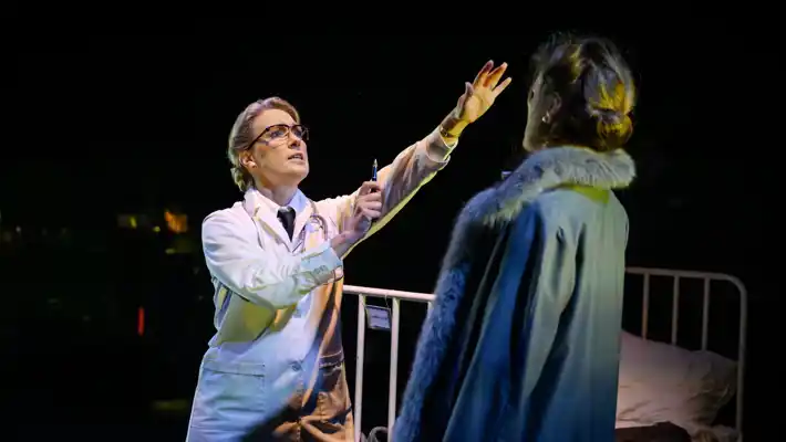 A woman in a doctor's coat extends her arm towards a woman in an overcoat, with a hospital bed in the background.