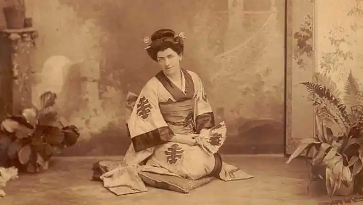 A woman sits, wearing a kimono, holding a fan in her hand.