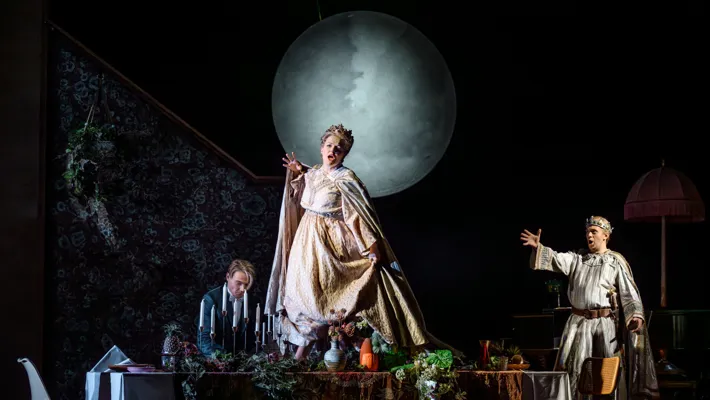 A woman stands on a set table wearing a crown, behind her a large moon and two men.