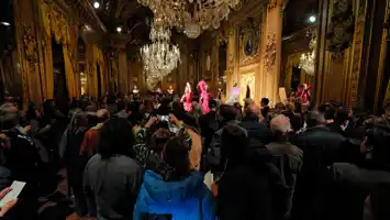 A large gold-decorated room with large crystal chandeliers: the room is completely full of people and two artists are standing on a stage.