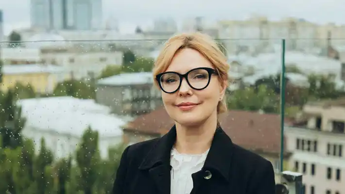 A young woman with glasses stands outdoors, a city spreads out in the background.