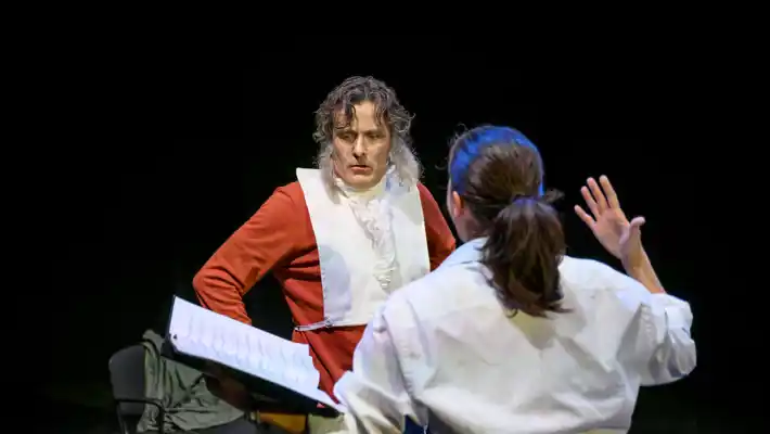 A man in red looks down at a sheet of music paper, in front of him a woman in a white shirt gesturing.